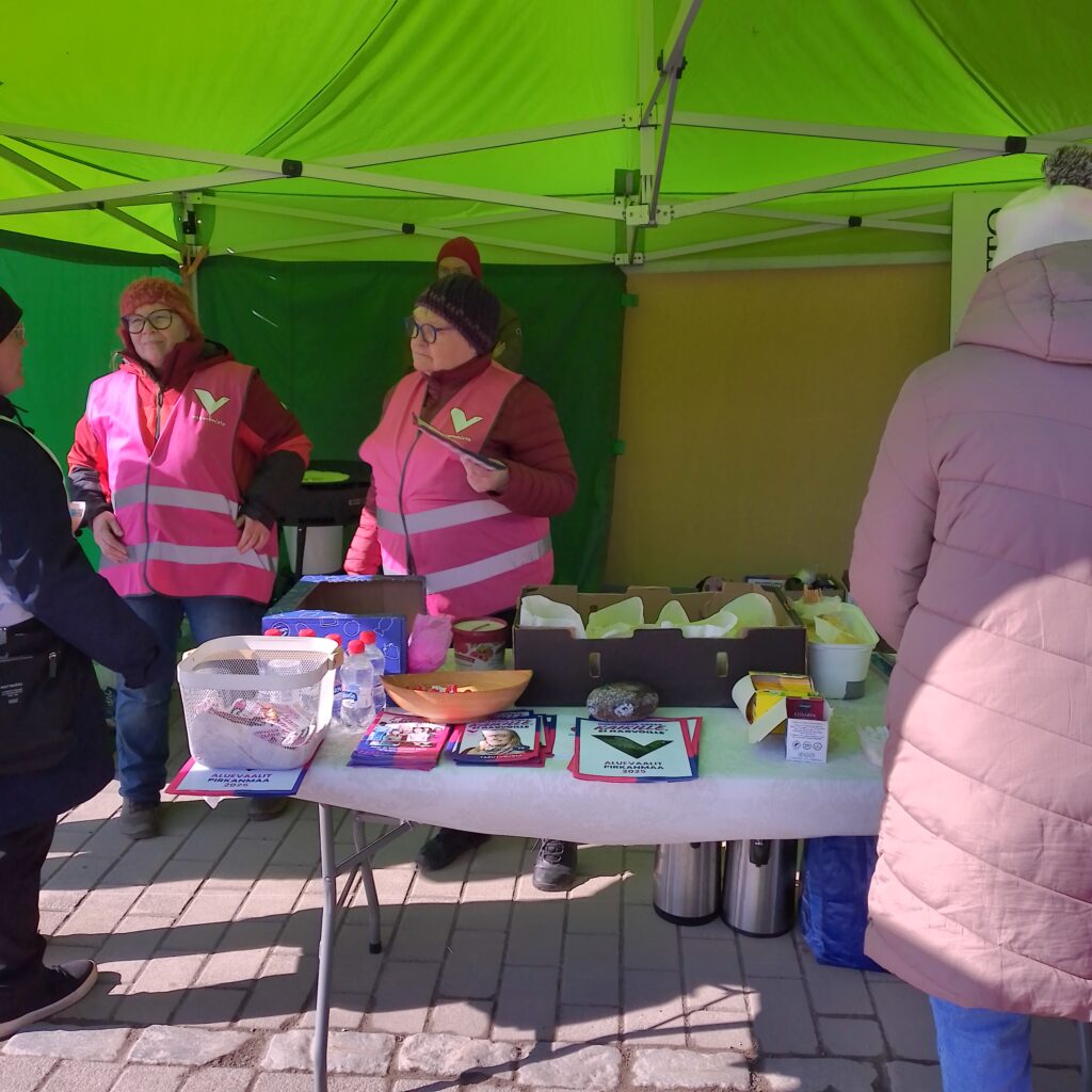 Sisällä vaaliteltassa. Etualalla pöytä, jossa vaalimateriaalia ja tarjoiluja. Pöydän takana seisovat Anne Kahelin ja Irma Koskela.